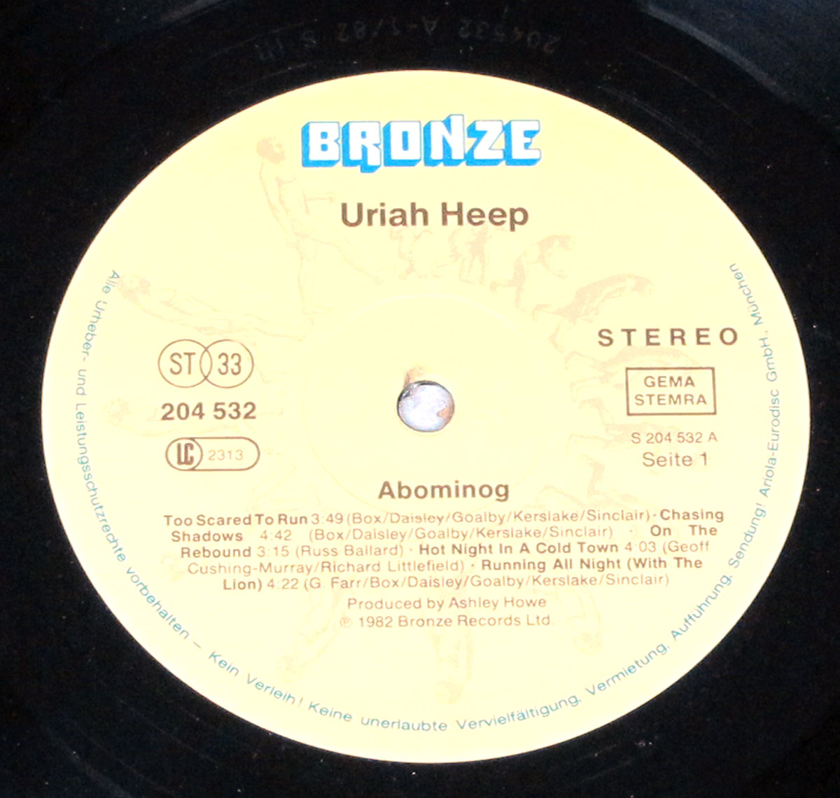Vinyl record label for Uriah Heep’s Abominog album. The label features a light beige background with the Bronze Records logo in blue at the top. Below it, the band’s name and album title are listed, along with the tracklist for Side 1. Standard record details, including catalog number, stereo designation, and copyright information, are included, with licensing text along the edge.